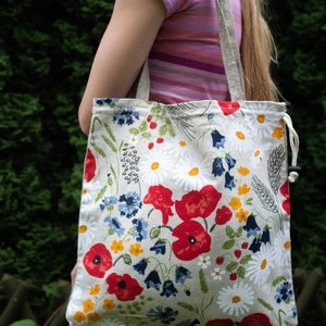 Può includere: Una grande borsa tote beige con stampa floreale con papaveri rossi, margherite bianche, campanule blu e foglie verdi. La borsa ha una chiusura con coulisse ed è realizzata in tessuto naturale.