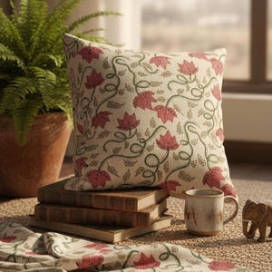 May include: A decorative pillow with a floral pattern in red and green on a cream background. The pillow is placed on a stack of books, with a matching napkin and a mug nearby. A potted fern is in the background.