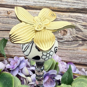 Puede incluir: Una estaca decorativa para jardín con una gran flor dorada en la parte superior de una esfera con estampado blanco y negro. La estaca tiene un tallo con cuentas y está rodeada de flores moradas y hojas verdes.