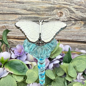 May include: A decorative bottle stopper with a silver butterfly design on a light blue glass base. The stem has blue and white accents. The stopper is surrounded by purple and white flowers and green leaves against a wooden background.
