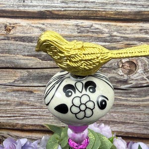 May include: A decorative bird figurine with a gold finish perched on a white and black floral patterned sphere. The sphere is connected to a purple beaded stem, creating a unique design. The background is a rustic wooden surface.