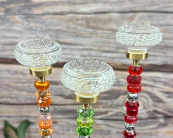 May include: Three decorative bottle stoppers with clear, etched glass tops and brass bases. The stems are adorned with colorful beads in shades of orange, green, and red. The background is a wooden surface.