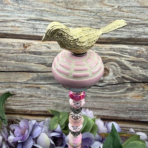 Puede incluir: Una estaca decorativa para jardín con un pájaro dorado posado sobre una pieza de cerámica rosa y esférica con un patrón de rejilla. La estaca está adornada con una columna de cuentas rosas, transparentes y plateadas. El fondo es una superficie de madera.