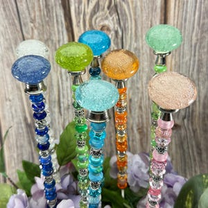 May include: A collection of colorful garden stakes with glass orb tops in shades of blue, green, orange, and pink. The stems are adorned with beads and silver accents, set against a wooden background and purple flowers.