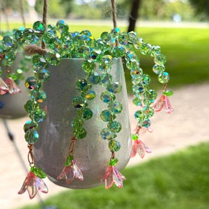 May include: A hanging planter featuring a speckled white ceramic pot with strands of faceted green crystal beads cascading over the sides. Each strand terminates in a small, translucent pink flower-shaped bead, suspended by twine.