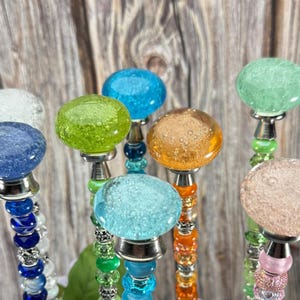 May include: A collection of decorative glass-topped garden stakes in various colors, including blue, green, orange, and pink. The stakes have beaded stems and silver-toned metal accents, perfect for adding a touch of color to any garden.