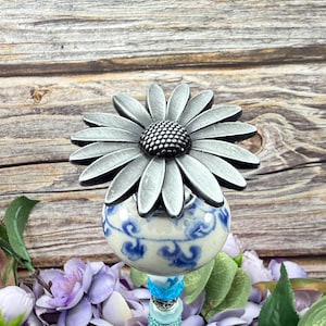 Op de afbeelding: Een decoratieve tuinsteker met een zilveren madeliefbloemkop op een blauw-witte keramische bol. De steker heeft een kralensteel in tinten blauw en turquoise. De achtergrond is een houten oppervlak met paarse bloemen en groene bladeren.