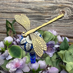 May include: A decorative dragonfly bottle stopper with gold-coloured wings and body. The stopper features a blue and white patterned ceramic bead and a blue glass stem. The dragonfly is set against a backdrop of purple and green flowers.