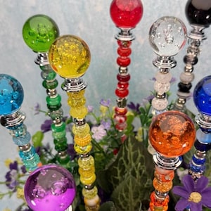 Puede incluir: Una colección de coloridos tapones decorativos para botellas. Cada tapón presenta un orbe de vidrio transparente y redondo en varios colores, como verde, amarillo, rojo y azul. Los orbes están montados sobre bases de metal plateado y adornados con tallos de cuentas.