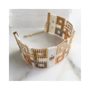 Pode incluir: Um bracelete de contas brancas com um padr&atilde;o geom&eacute;trico em tons de azul, laranja e dourado. O bracelete tem um fecho dourado.
