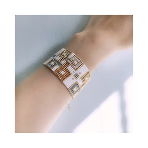 Pode incluir: Pulseira de contas brancas com um padr&atilde;o geom&eacute;trico em dourado e azul. A pulseira &eacute; feita com contas pequenas e tem um fecho de corrente delicado.