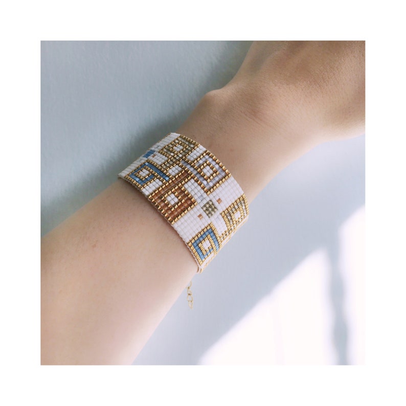 Pode incluir: Pulseira de contas brancas com um padr&atilde;o geom&eacute;trico em dourado e azul. A pulseira &eacute; feita com contas pequenas e tem um fecho de corrente delicado.