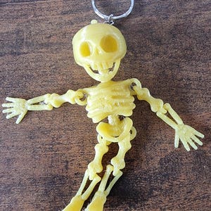 May include: A yellow skeleton keyring with a silver ring. The skeleton has articulated arms and legs, and a smiling skull. The keyring is made of plastic and is designed to be a fun accessory.