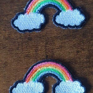 May include: Two embroidered rainbow patches. Each patch features a colorful rainbow arching over a white cloud with a black outline. The patches are likely for clothing or accessories.