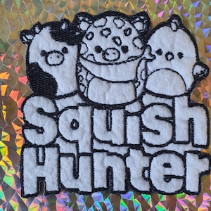 Può includere: Una toppa bianca con tre personaggi di animali dei cartoni animati e la scritta "Squish Hunter" in nero. Gli animali sono una mucca, un maiale e un uccello. La toppa è su uno sfondo olografico.