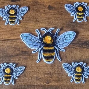 May include: Five embroidered bee patches. Each bee features a yellow head and abdomen with black stripes. The wings are light blue with detailed stitching. The patches are on a brown wooden surface.