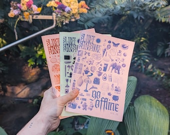May include: A hand holds three journals with the words "go offline" and various illustrations. The journals are in pink, green, and orange. The background features lush green foliage and a floral arrangement.