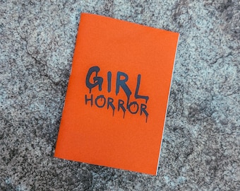 May include: An orange notebook with the words "GIRL HORROR" in black, stylized lettering. The notebook is resting on a textured, grey stone surface.