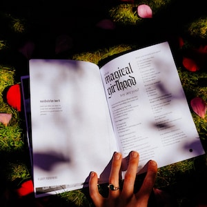 May include: An open book with the title "What magical girlhood looks like to you?" The book is lying on a bed of green grass with pink flower petals scattered around it.