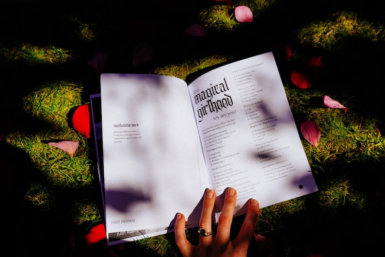 May include: An open book with the title "What magical girlhood looks like to you?" The book is lying on a bed of green grass with pink flower petals scattered around it.