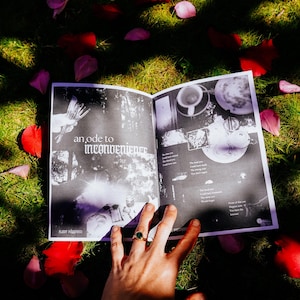May include: An open book with black and white pages. The book is lying on a bed of green grass and pink flower petals. The text on the page reads "an ode to inconvenience".