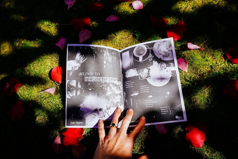 May include: An open book with black and white pages. The book is lying on a bed of green grass and pink flower petals. The text on the page reads "an ode to inconvenience".