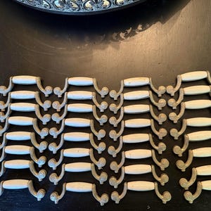 Könnte beinhalten: Eine Sammlung von Vintage-Schrankgriffen mit cremefarbenem Mittelteil und aufwendigen, gealterten Metalldetails. Die Griffe sind in Reihen auf einer dunklen Oberfläche angeordnet, was auf ein Hardware- oder Wohnkulturthema hindeutet.