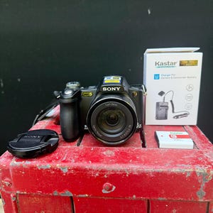 May include: A black Sony digital camera with a lens cap, a charger box, and a battery pack are on a red surface. The camera has a Carl Zeiss lens and the text "SONY" is visible on the camera.
