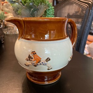 May include: A brown and white ceramic pitcher with a brown handle. The pitcher has a white design of a person riding a horse with blue polka dots.