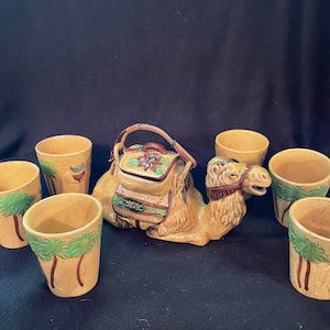 May include: A ceramic camel teapot and six matching cups. The camel is brown and tan with a brown saddle and green palm trees on the cups.