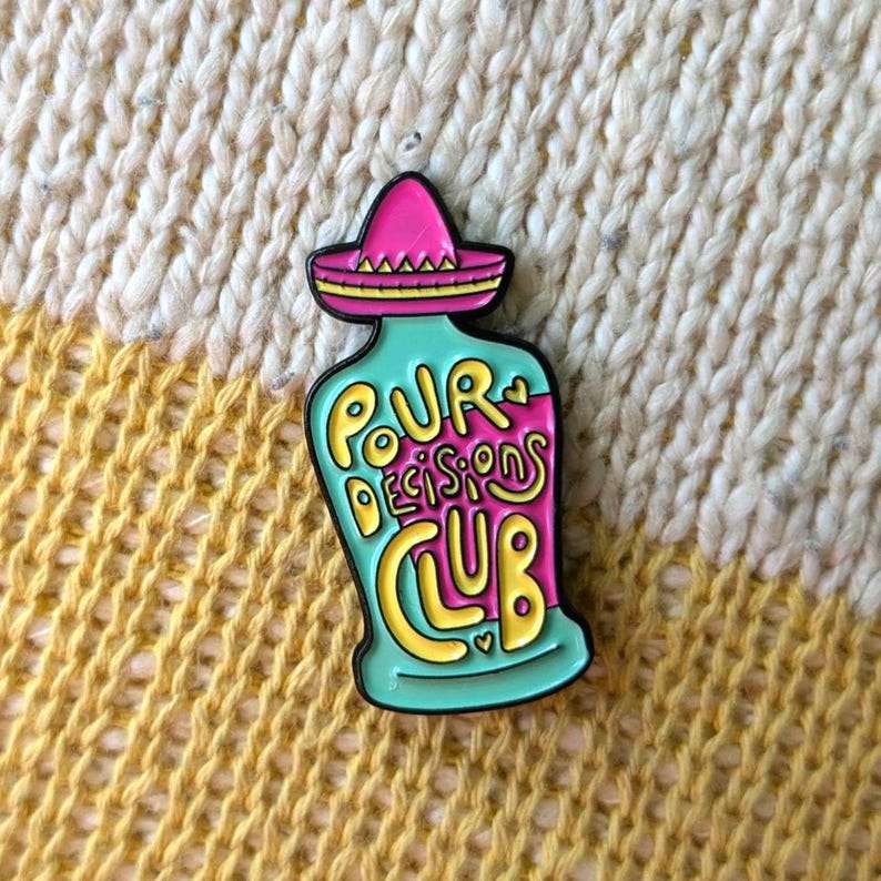 May include: A teal blue enamel pin shaped like a bottle with a pink sombrero on top. The bottle has the words "Pour Decisions Club" written on it in yellow and pink.