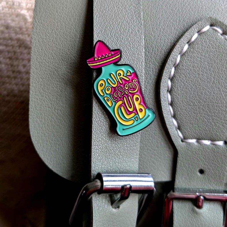 May include: A teal blue enamel pin shaped like a bottle with a pink sombrero on top. The bottle has yellow lettering that says "Pour Decisions Club".
