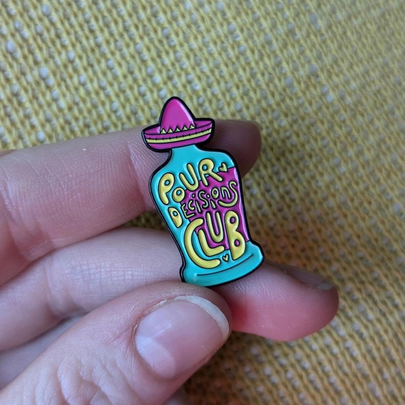 May include: A turquoise blue enamel pin shaped like a bottle with a pink sombrero on top. The bottle says "Pour Decisions Club" in yellow and pink lettering.
