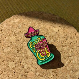 May include: A green and pink enamel pin shaped like a bottle with a sombrero on top. The bottle says "Pour Decisions Club" in yellow lettering.