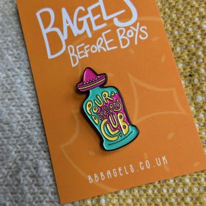 May include: A teal blue enamel pin with a pink sombrero on top. The pin depicts a bottle with the text "Pour Decisions Club" on it. The pin is on a bright orange background with the text "Bagels Before Boys" and "bbbagels.co.uk".