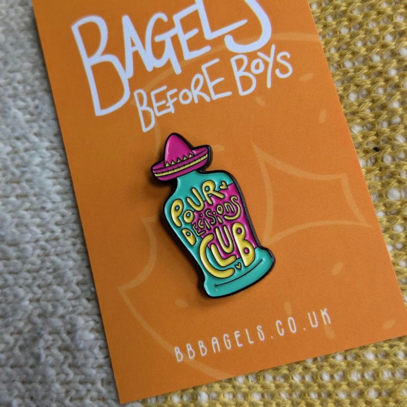 May include: A teal blue enamel pin with a pink sombrero on top. The pin depicts a bottle with the text "Pour Decisions Club" on it. The pin is on a bright orange background with the text "Bagels Before Boys" and "bbbagels.co.uk".
