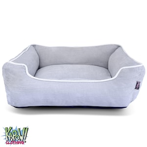 Puede incluir: Una cama rectangular para mascotas con un estampado de rayas grises y blancas. La cama tiene una base azul marino y ribetes blancos. El interior es de color gris claro liso. La cama tiene una etiqueta que dice "PeeWee".