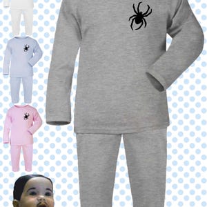 May include: Gray pajama set with a black spider graphic on the chest. The long-sleeved top and pants are made of a soft material. Additional pajama sets in white, light blue, and pink are also shown.