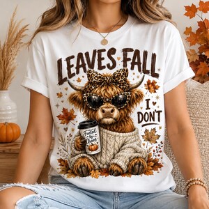 May include: A white t-shirt featuring a graphic of a Highland cow wearing a leopard print bow and sunglasses, dressed in a beige cable-knit sweater. The cow holds a coffee cup labeled "Fueled by Chaos" with the text "LEAVES FALL I DON'T" around it.