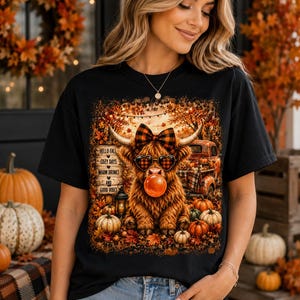 Könnte beinhalten: Ein schwarzes T-Shirt mit einer Grafik eines flauschigen Hochlandrindes, das eine karierte Schleife und herzförmige Sonnenbrillen trägt und eine Blase bläst. Das Design enthält ein Holzschild mit dem Text Hello Fall, Cozy Days, Warm Drinks and Good Vibes.