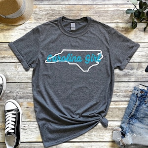 May include: A gray t-shirt with a white outline of the state of North Carolina and the text "Carolina Girl" in blue.
