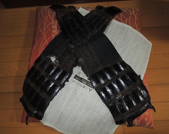 May include: Pair of black, segmented armor gauntlets, likely for the arms, with a dark brown trim. The gauntlets are resting on a red and white patterned cushion, with a newspaper underneath. The armor appears to be made of leather or a similar material.