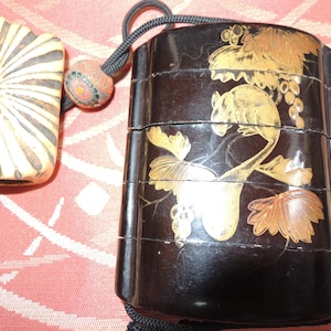 Op de afbeelding: Een zwarte, gelaagde Japanse bento-box met gouden bloemmotieven, een gesneden ivoorkleurige stopper en een decoratieve kraal. De bento-box heeft een zwart koord bevestigd. De stopper heeft een radiaal ontwerp.