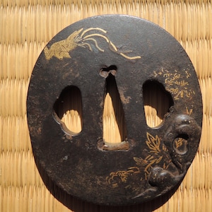 May include: A circular, dark metal Japanese tsuba, or sword guard, featuring gold-coloured floral and bird designs. The tsuba has three openings and is set against a woven straw background.