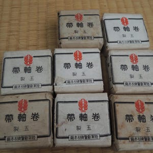 May include: Eight small, rectangular packages are arranged on a woven mat. Each package is wrapped in off-white paper and features a black border with Japanese text and a red emblem. The packages are small and square.