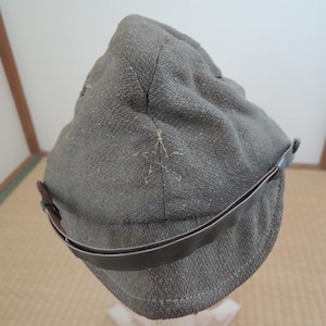 May include: A gray, textured cap with a dark brown leather visor and strap. The cap has a star-shaped embroidered detail on the side. The cap is made of a woven fabric.