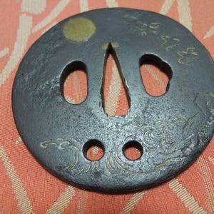 Op de afbeelding: Een donkergrijs, cirkelvormig metalen object met verschillende gaten en ingewikkelde geëtste ontwerpen. Een goudkleurige cirkel bevindt zich bovenaan. Het object heeft een centrale opening en is waarschijnlijk een decoratief of functioneel stuk.