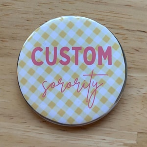 Può includere: Un bottone rotondo con un motivo a quadri giallo e bianco. Il bottone presenta le parole "CUSTOM" e "sorority" in corsivo rosa. Il bottone è su una superficie di legno.