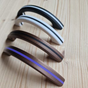 Pode incluir: Quatro puxadores de armário em fila, cada um com um design curvo. Os puxadores são pretos com uma faixa branca, brancos com uma faixa preta, castanhos com uma faixa preta e castanhos com uma faixa roxa. Sobre uma superfície de madeira clara.