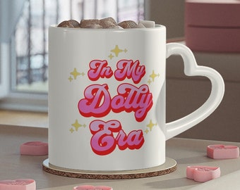 May include: White ceramic mug with a heart-shaped handle, filled with a dark substance, and the text "In My Dolly Era" in pink and red. The mug sits on a wooden coaster, surrounded by pink heart-shaped candies.